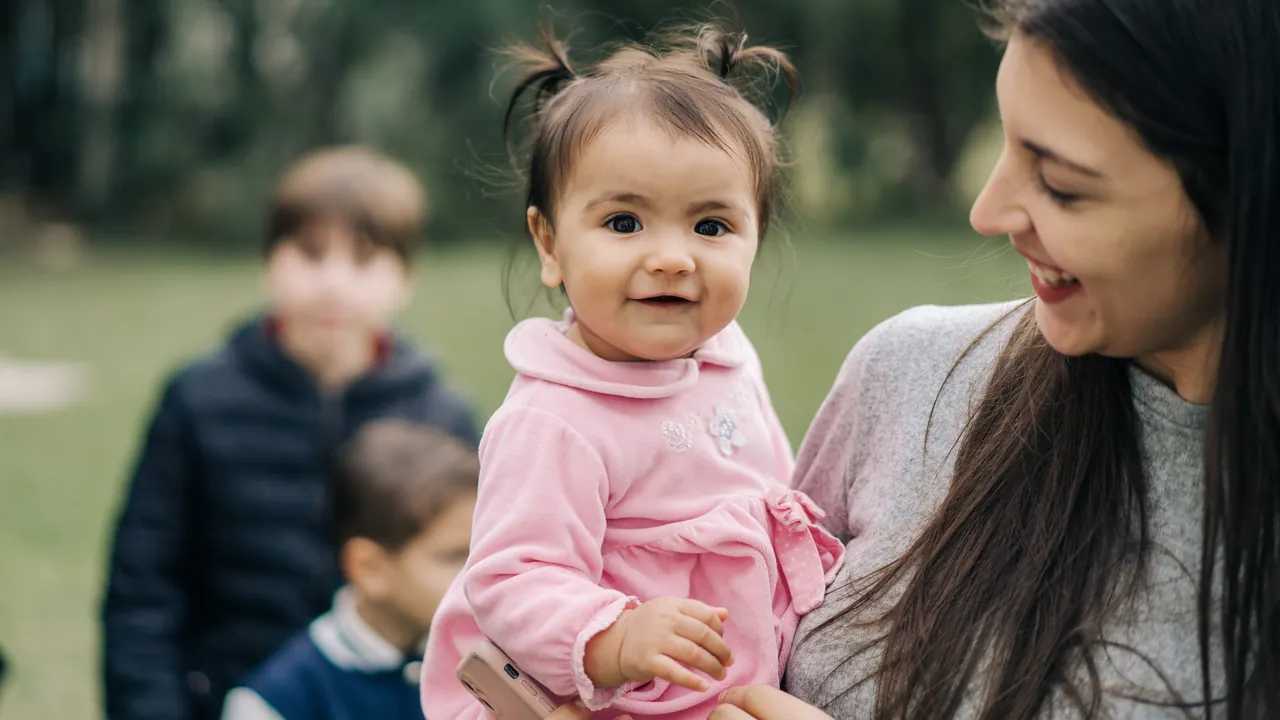 Mariana Gouvêa segurando um bebê no colo ao ar livre, com duas crianças ao fundo