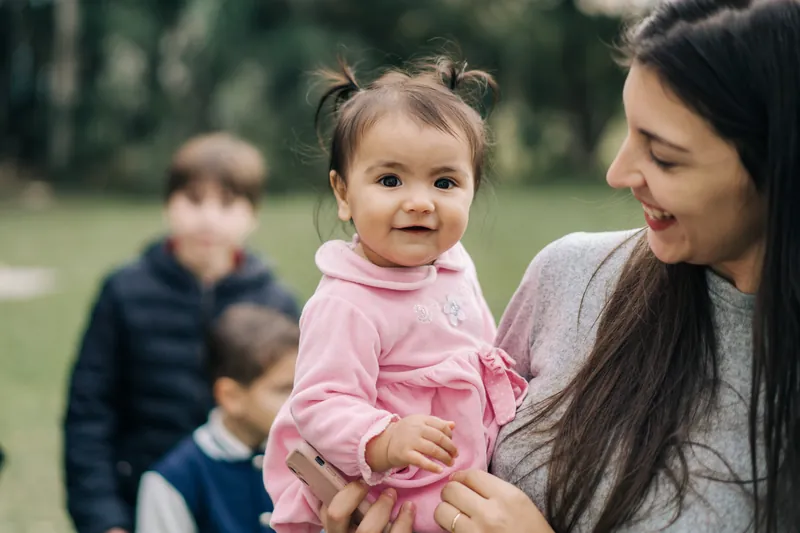 Mariana Gouvêa segurando um bebê no colo ao ar livre, com duas crianças ao fundo