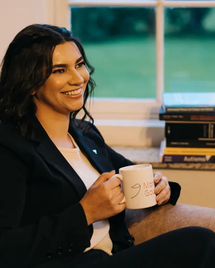 Mariana Gouvêa sentada em poltrona, sorrindo com uma xícara da marca, livros ao fundo